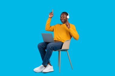 Modern dizüstü bilgisayar ve kablosuz kulaklık kullanarak mavi stüdyo arka planında izole edilmiş bir sandalyede oturmuş müzik dinleyen, kopyalama alanı, tam boy...