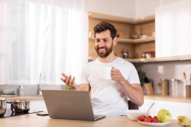 Beyaz tişörtlü, gülümseyen, beyaz, beyaz sakallı, modern mutfak içinde el kol hareketi yapan bir beyaz erkek. İş, evle uzaktan iletişim, online ders ve sosyal mesafe toplantısı.