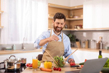 Önlüklü, sakallı, mutlu yetişkin beyaz bir erkek yemek blogu için video çekiyor. Modern mutfak mutfağında organik sebzelerle tuzlu salata yapıyor. Ev yapımı yemek, yeni tarif, ev yapımı cihaz.