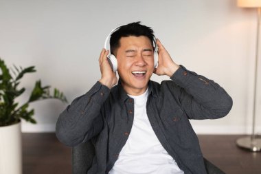 Orta yaşlı, kablosuz kulaklıklı, gözleri kapalı Asyalı bir erkek müzik dinliyor, boş zamanın tadını çıkarıyor, oturma odasının iç kısmında koltukta oturuyor, kapat. Modern aygıtın ses uygulaması, covid-19