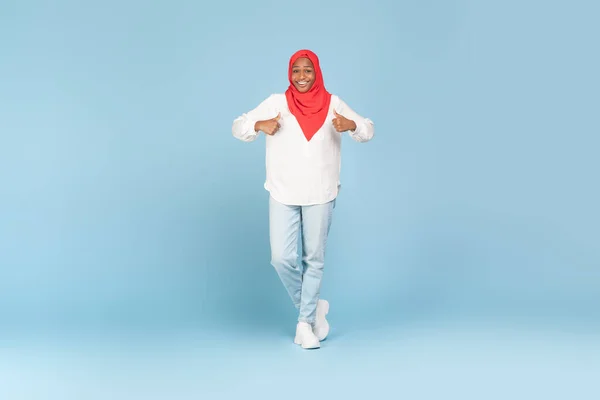 Positive african american muslim woman showing thumbs up and smiling at camera on blue studio background, full length shot with copy space for advertisement