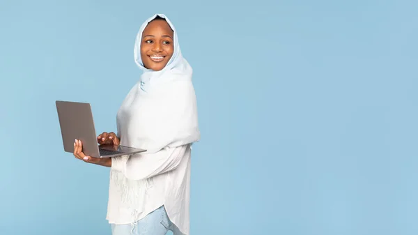 穿着头巾笑着的黑人女人 站在笔记本电脑旁边 看着蓝色工作室背景上的空旷空间 学习或使用笔记本的非裔美国穆斯林女士 — 图库照片