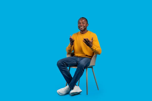 宽松的中年黑人男子穿着休闲装坐在椅子上摆姿势 在蓝色工作室的背景上对着相机微笑 拍摄全长镜头 复制广告空间 — 图库照片