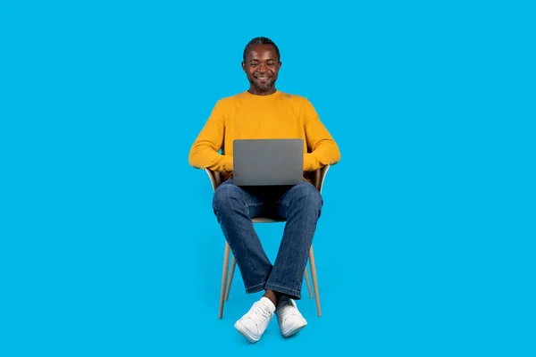 穿着黄色毛衣坐在椅子上的快乐的非洲中年男人 在蓝色的工作室背景上使用现代笔记本电脑 在笔记本电脑键盘上打字 上网工作或玩游戏 复制空间 — 图库照片