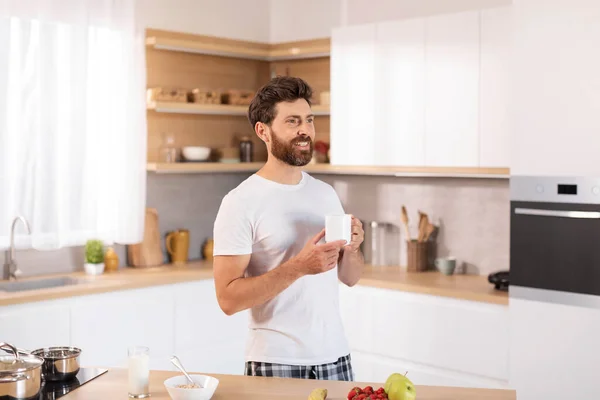 快乐的中年白人男性 留着胡子 穿着白色T恤 喝着热饮 看着现代厨房内部空旷的空间 在家喝咖啡 早上和早餐 伟大的广告和提供 — 图库照片