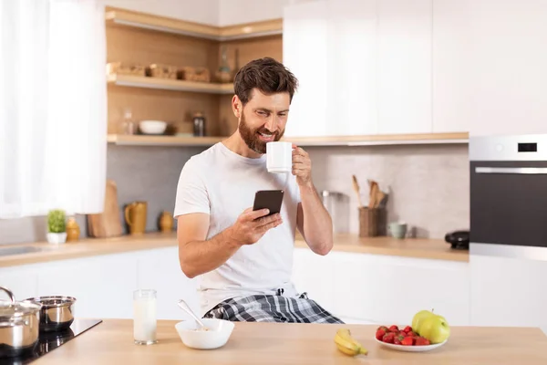 快乐成熟的高加索男人 留着胡子 穿着白色T恤 喝着杯子 在厨房的内部用电话打字 博客和社交媒体应用 在家喝咖啡休息 早上好 广告和服务 — 图库照片