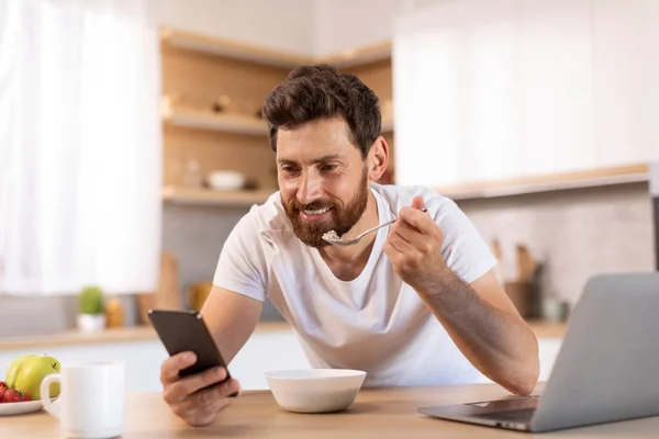 身穿白色T恤 留着胡子的成熟的高加索男性微笑着吃粥 在智能手机上阅读新闻 在现代厨房内部观看笔记本电脑上的视频 早上好 在家工作的应用程序 — 图库照片