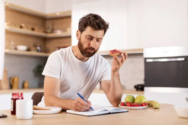 忙碌而集中的成年白人男性 留着胡子 穿着白色T恤 吃三明治 在现代厨房内部的笔记本上记笔记 早上和早餐单独在家里 计划和工作 — 图库照片