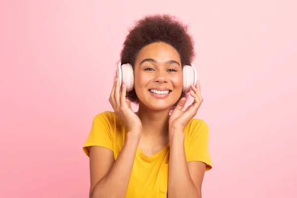 stock image Smiling millennial african american curly female student in casual and wireless headphones, enjoy music, isolated on pink background, studio. Audio app and device for relaxing in free time, emotions