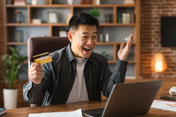 喜形于色的中国男性兴奋地张开嘴 拿着信用卡 在家里办公室的桌子上看着电脑 成功的工作 遥远的商业 金融和感情 — 图库照片