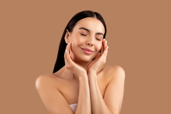 Skin care cosmetics. Sensual half-naked young woman with perfect body touching her face with closed eyes, enjoying silky smooth skin after face care routine, isolated on beige studio background