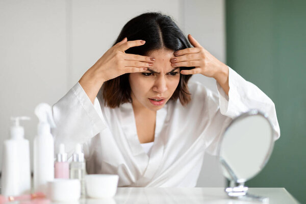 Angry middle eastern young woman in white silky robe sitting at vanity table full of beauty products, looking at mirror, checking skin, touching her forehead, home interior. Acne, pimples, dull skin