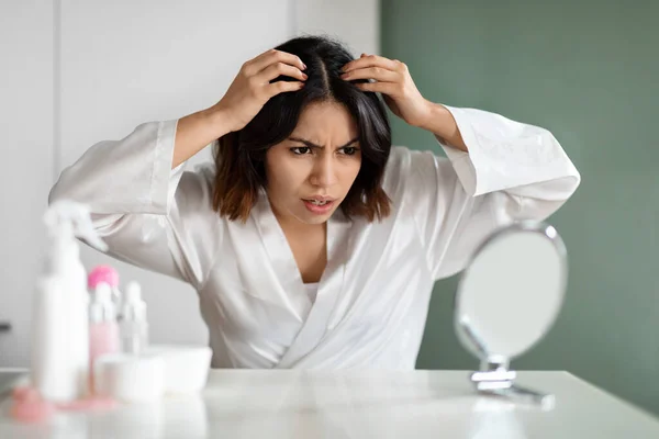Angry young arab lady in white bathrobe sitting in bedroom, checking her hair roots, looking at mirror, having disappointed face expression, copy space. Hair thinning, hair loss, grey hair concept
