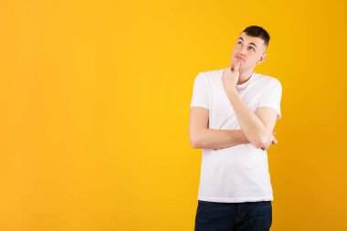 Düşünceli beyaz adam fotokopi odasına bakıyor ve çenesine dokunuyor. İlginç bir teklif düşünüyor. Sarı stüdyo arka planında duruyor.