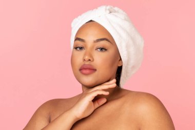 Güzellik konsepti. Afro-Amerikan tombul kadının portresi. Kişisel bakım sırasında kafasında havluyla poz veriyor. Pembe arka plan. Spa ve sağlık