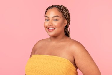 Güzel Afro-Amerikan tombul kadın portresi, kusursuz pürüzsüz tenli banyo havlusuna sarılmış, kameraya bakıyor ve gülümsüyor, pembe stüdyo arka planında poz veriyor.