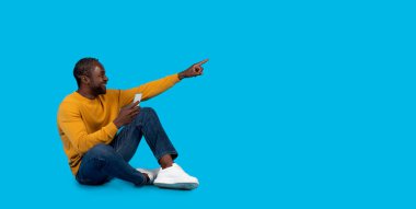 Güzel online teklif. Gülümseyen orta yaşlı, şık günlük kıyafetli bir zenci akıllı telefonuyla yerde oturuyor ve mavi stüdyo arka planındaki reklam için fotokopi alanını işaret ediyor.