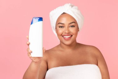 Mutlu Afro-Amerikan tombul kadın pembe arka planda boş şampuan şişesi, reklam vücudu ve saç bakım ürünleri gösteriyor. Kozmetik ürün reklamı için bir model