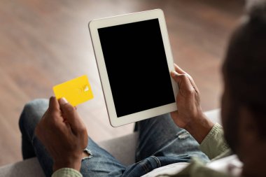 Kanepede oturan Afro-Amerikan bir adam boş ekranlı bir tabletle video izliyor, kredi kartını odanın içinde gösteriyor. Para, alışveriş kolik evde online alışveriş, büyük indirim, bankacılık ve finans uygulaması