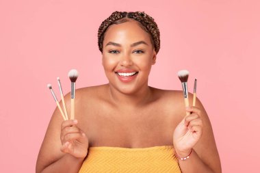 Uydurma konsept. Makyaj fırçaları tutan, pembe stüdyo arka planında havluya sarılı duran, siyah, güzel bir kadının portresi.