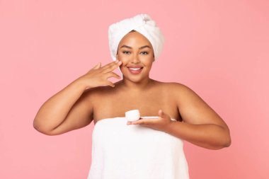 Günlük cilt bakımı. Elinde krema kavanozu tutan, yanağına nemlendirici süren ve kameraya poz veren şişman Afrikalı Amerikalı kadının portresi. Kafasına beyaz havlu geçirmiş siyah tombul bir kadın.