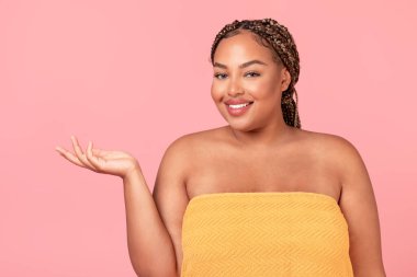 Güzellik reklamı. Mutlu siyah beden pozitifliği olan kadın elinde boş avuçla pembe arka planda reklam için fotokopi alanı gösteriyor.