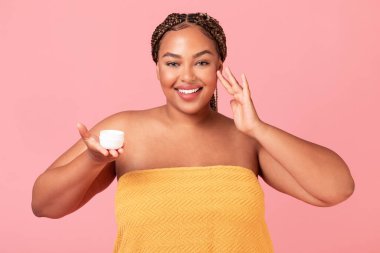 Mutlu siyah obez kadın yüzüne nemlendirici krem sürüyor, havluya sarılmış vaziyette duruyor, kozmetik ürünlerle dolu kavanozu tutuyor ve kameraya gülümsüyor.