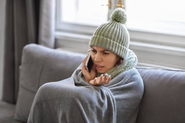 Donmuş genç bir kadın battaniyeye sarılmış evde telefon görüşmesi yapıyor, koltukta oturan ve cep telefonuyla konuşan stresli bir kadın apartman dairesinde soğuk hava koşullarından yakınıyor,