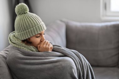 Evde kanepede otururken elleri ısıtan, donan kadın örgü ören kadın, içeride üşüyen eşarp giyen, battaniyeye sarılı, sıcacık ısıtma problemleri yaşayan kadın.