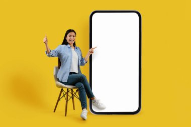 Bin yıllık Çinli kız öğrencinin gündelik sandalyede oturmasına sevindim. Sarı arka planda boş ekranı olan büyük akıllı telefonu işaret etmesine sevindim. Yeni uygulamanın sunumu, büyük indirim, reklam ve teklif