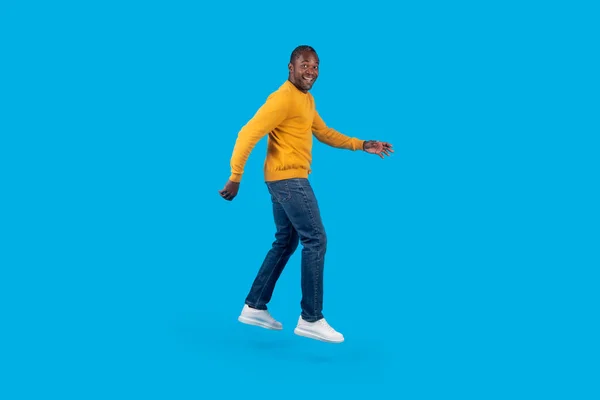 感情的な幸せな魅力的な成熟したアフリカ系アメリカ人男性でスタイリッシュなカジュアルな衣装でジャンプアップ上の青いスタジオの背景 実行しているか 空気中で踊る フル長さ 広告のためのコピースペース — ストック写真