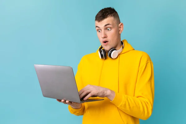 Emotional caucasian guy looking at laptop screen in shock, excited about win or success over blue background. Young man with portable pc cannot believe huge online sale