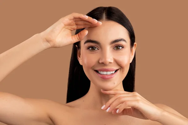 Skin care routine concept. Headshot of happy half-naked millennial woman touching her face and smiling, demonstrating silky smooth glowing young skin after face care routine, beige studio background