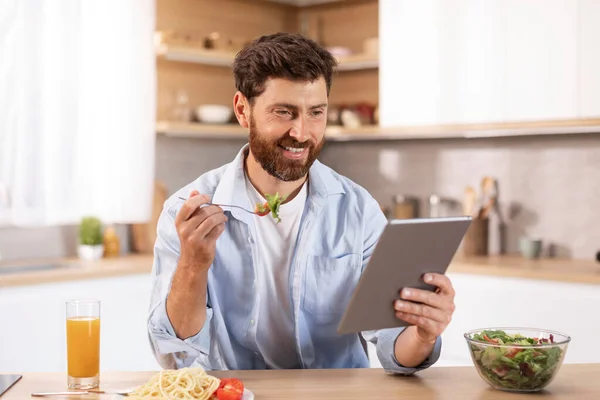 令人高兴的是 有胡子的高加索成年人吃午饭 在平板电脑上阅读新闻 在现代厨房的室内观看视频 与社交距离的学习 家里的设备 应用程序 极好的建议和广告 — 图库照片