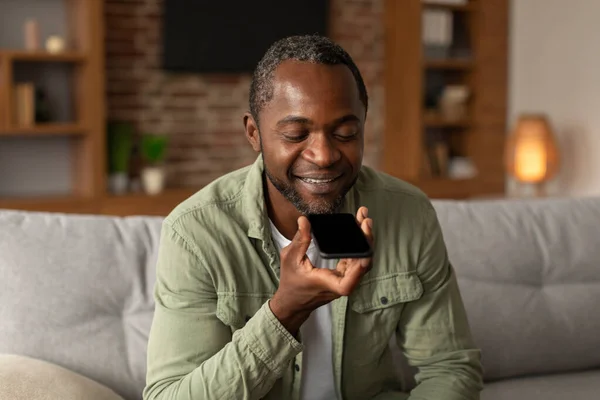 ソファの上でアフリカ系アメリカ人の中年男性の笑顔は スマートフォンで白い画面で呼び出し 話して リビングルームのインテリアでチャットします リモートで通信するための新しいアプリ 自宅でガジェットや良いニュース — ストック写真
