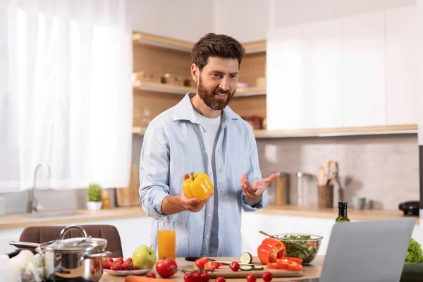 Glad mature caucasian guy with beard has video call, makes video at laptop, holds pepper in hands, prepares eat in modern kitchen interior. Homemade food, health care, meal blog with device at home