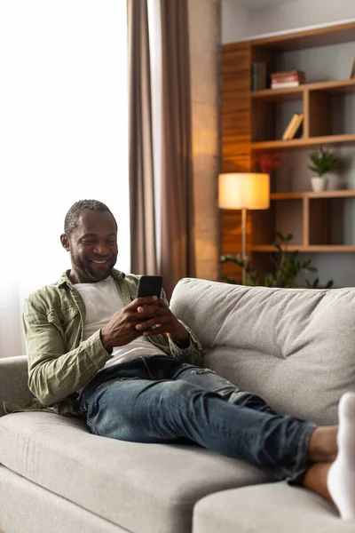 笑着在网上冲浪的中年非洲裔美国人 在智能手机上读到信息 在客厅里免费观看视频 用于社交网络 搜索和新家用电器的应用程序 — 图库照片