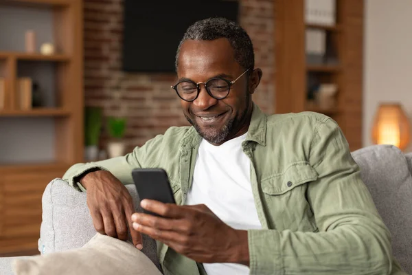 中年アフリカ系アメリカ人男性の笑顔がスマートフォンでメッセージを読み取り リビングルームのインテリアでビデオを視聴します ソーシャルネットワークのためのアプリ ブログ インターネットでサーフィン 自宅で新しいデバイス — ストック写真