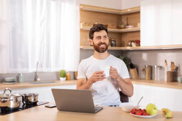Glad adult caucasian guy with beard in white t-shirt enjoys cup of coffee, sits at table with laptop, thinks, looks at free space in modern kitchen interior. Work, business at home, idea for startup