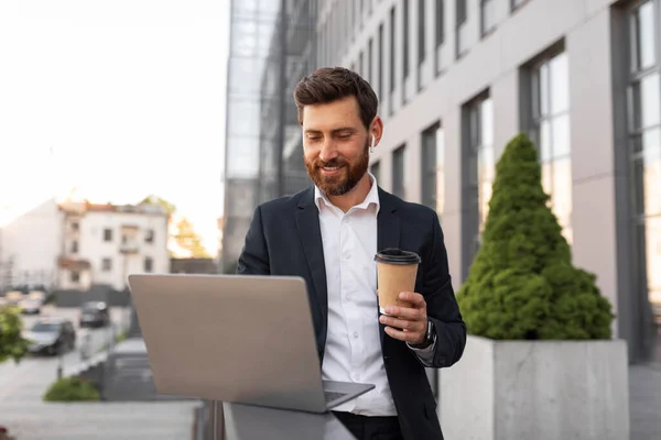 Happy confident young european man boss with beard in suit, wireless headphones typing on computer with cup of drink takeaway on balcony of building. Coffee break, app for work, finance and business