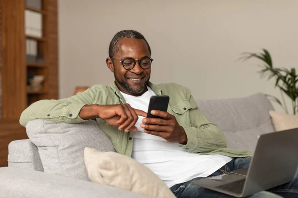 中世のアフリカ系アメリカ人の男性が電話でメッセージを読み取り 室内でビデオサーフィンを見て スペースをコピーします ソーシャルネットワークのためのアプリ ブログ 自宅でインターネットや新しいデバイスでの検索 — ストック写真