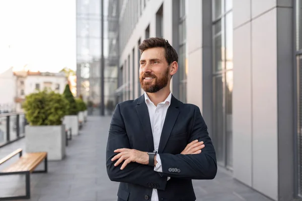 Glad confident handsome young european male ceo manager or general director with beard in suit looks at empty space near building on city street. Modern business, career, leadership, ad and offer