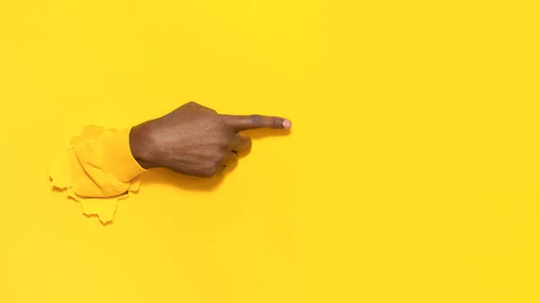 African american male hand pointing aside on text or your design with index finger, gesturing through the torn yellow paper background, closeup, free space