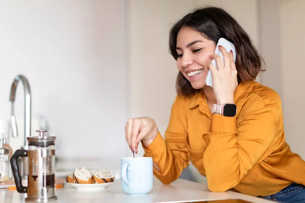 stock image Happy Young Arab Woman Drinking Coffee And Talking On Mobile Phone In Kitchen, Smiling Middle Eastern Female Stirring Sugar In Cup And Enjoying Pleasant Cellphone Conversation, Closeup Portrait