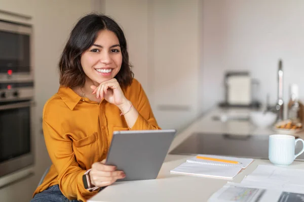 Portrait Of Smiling Young Arab Female Freelancer Using Digital Tablet While Sitting At Kitchen Counter, Happy Middle Eastern Woman Working With Modern Gadget And Papers At Home, Copy Space