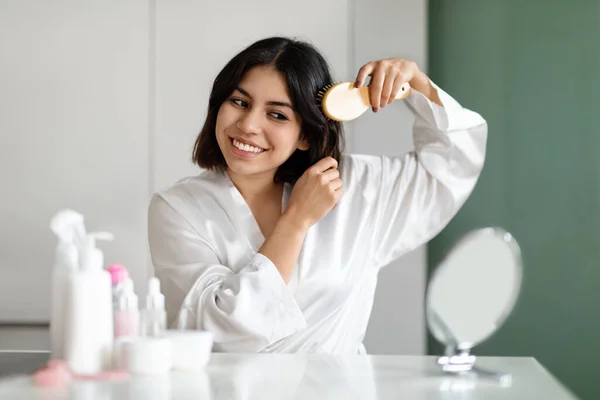 Beauty Routine. Pretty Middle Eastern Woman Combing Her Beautiful Short Hair With Brush In Front Of Mirror At Home, Attractive Young Lady Looking At Her Reflection And Smiling, Copy Space
