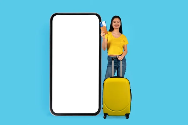 Smiling millennial japanese lady blogger with suitcase shows passport and tickets, near huge smartphone with empty screen, isolated on blue background, studio. App for travel order, vacation and ad