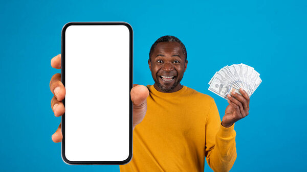 Excited emotional african american middle aged man in yellow gambling online, showing brand new cell phone with white blank screen and bunch of cash dollars on blue, recommending nice app, mockup
