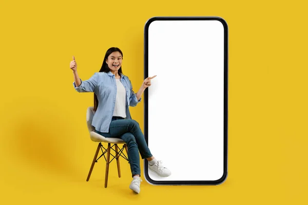 欣慰的是 千禧年的中国女学生坐在椅子上 手指指向大智能手机 屏幕空旷 背景是黄色的 新应用的展示 巨大的销售 广告和提供 — 图库照片