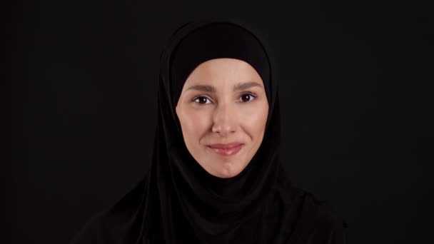 特写年轻的穆斯林女子 头戴头巾 脸朝下对着相机 面带微笑地对着相机 背景是黑色的 动作缓慢 — 图库视频影像
